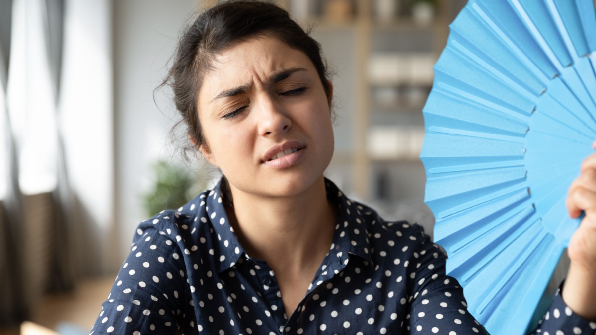 Overheated indian woman sit at workplace without air conditioner hold blue handheld fan cools herself frown face closed eyes feels unwell. Too hot summer weather day, high temperature indoors concept.