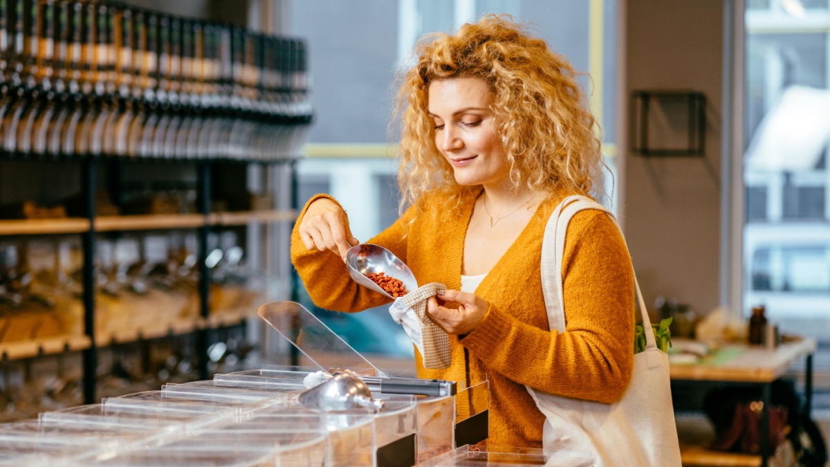 Sustainable shopping at small local businesses. Blond curly female in yellow cardigan putting Goji berries in cotton produce bag at plastic free store. Reusable eco bag for shopping.Zero waste concept