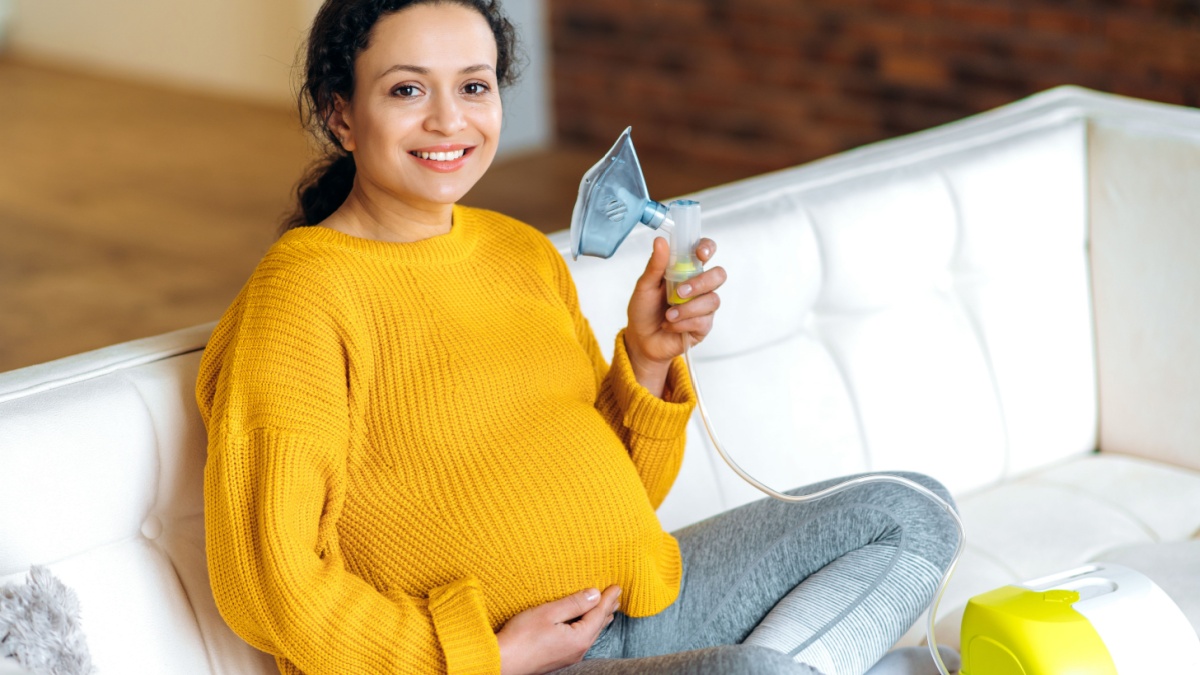 Joyful recovering pregnant mixed race brunette woman, sitting in the living room on the couch wearing a yellow sweater, does preventive treatment, holds an inhaler in her hand, looking at the camera.