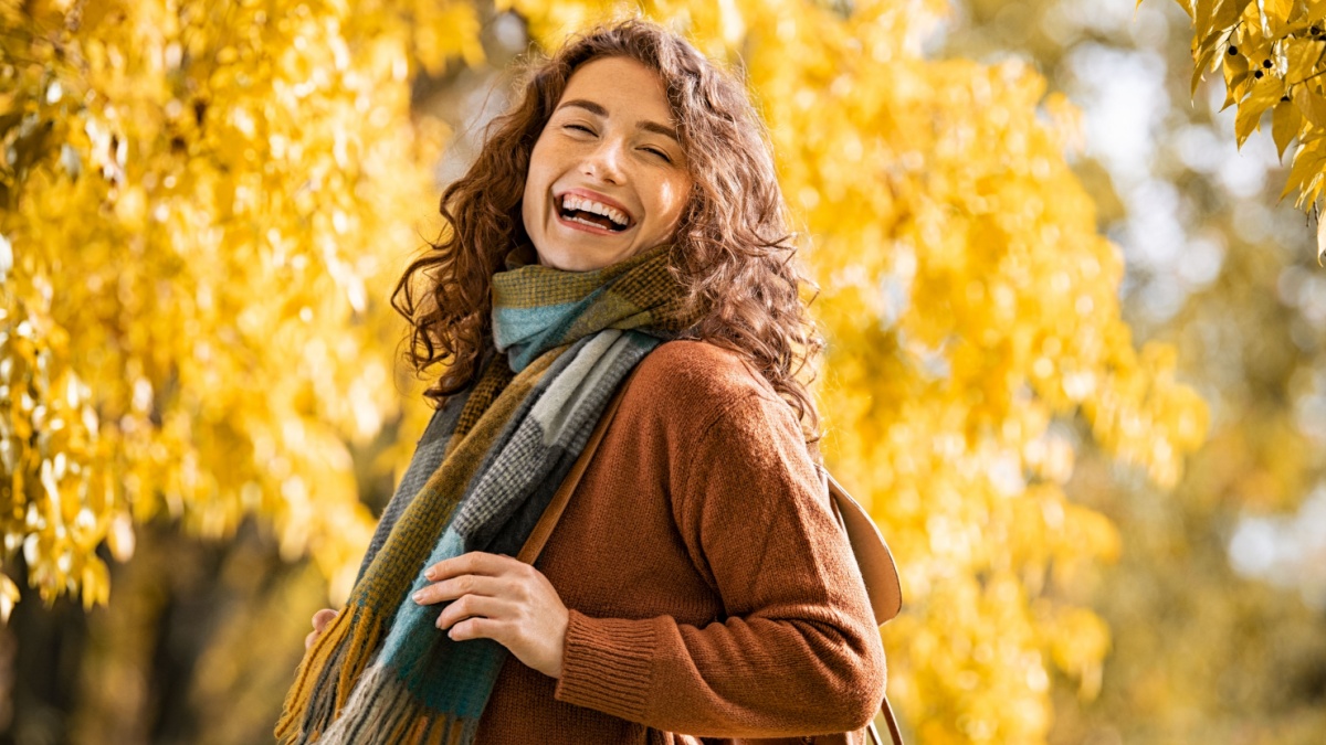 Carefree young woman wearing scarf with a big grin on face at park during autumn season. Smiling casual girl with warm and winter clothes enjoying sunny day at park with yellow trees on background.