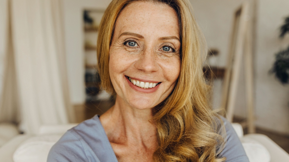 Senior woman-blogger with long golden hair, pretty freckled face, blue eyes and charming smile making selfie or recording stories for social network page, looking friendly and happy.