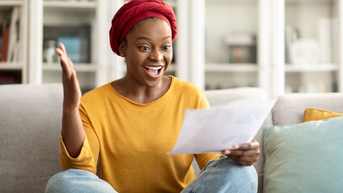 Happy young african american woman sit on couch at home, reading good news in paper letter checking domestic bills, emotional black lady holding documents doing paperwork, reading correspondence.