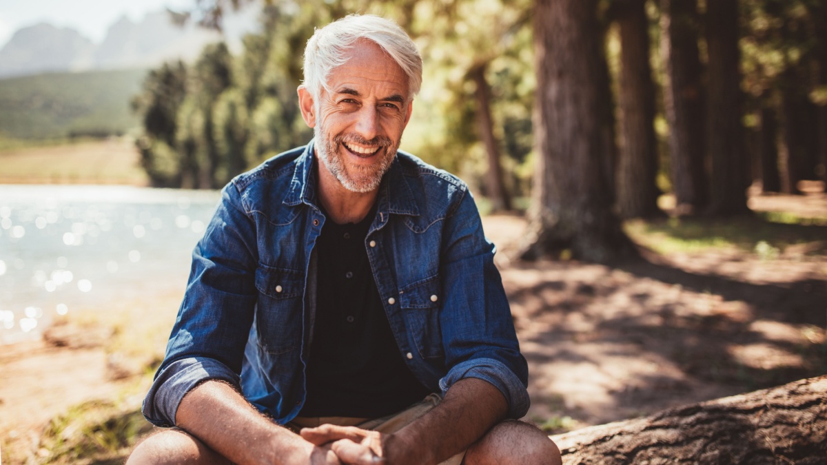 Portrait of happy mature man sitting near a lake looking at camera and smiling. Senior caucasian man sitting on a log by the lake on a summer day.