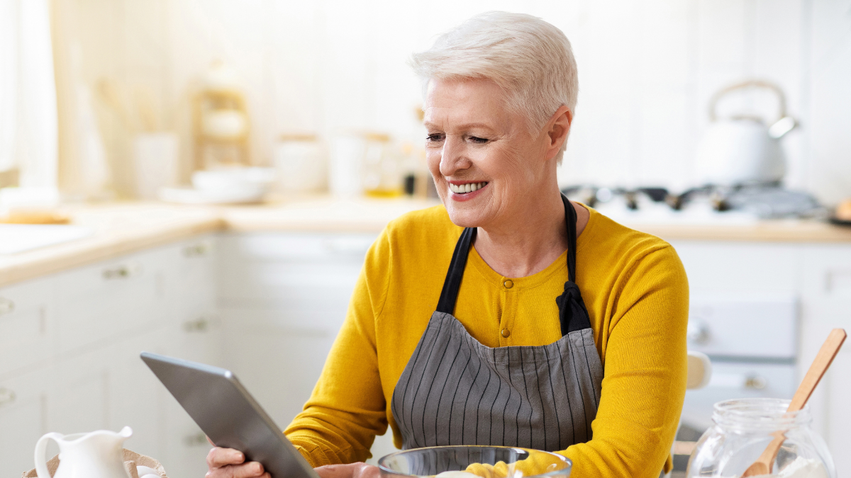 Cheerful aged lady cooking in kitchen, holding digital tablet doing meal prep.