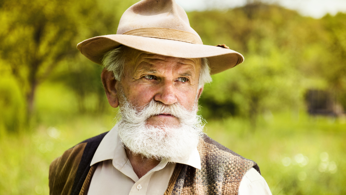 Portrait of old farmer with beard and hat in his backyard.