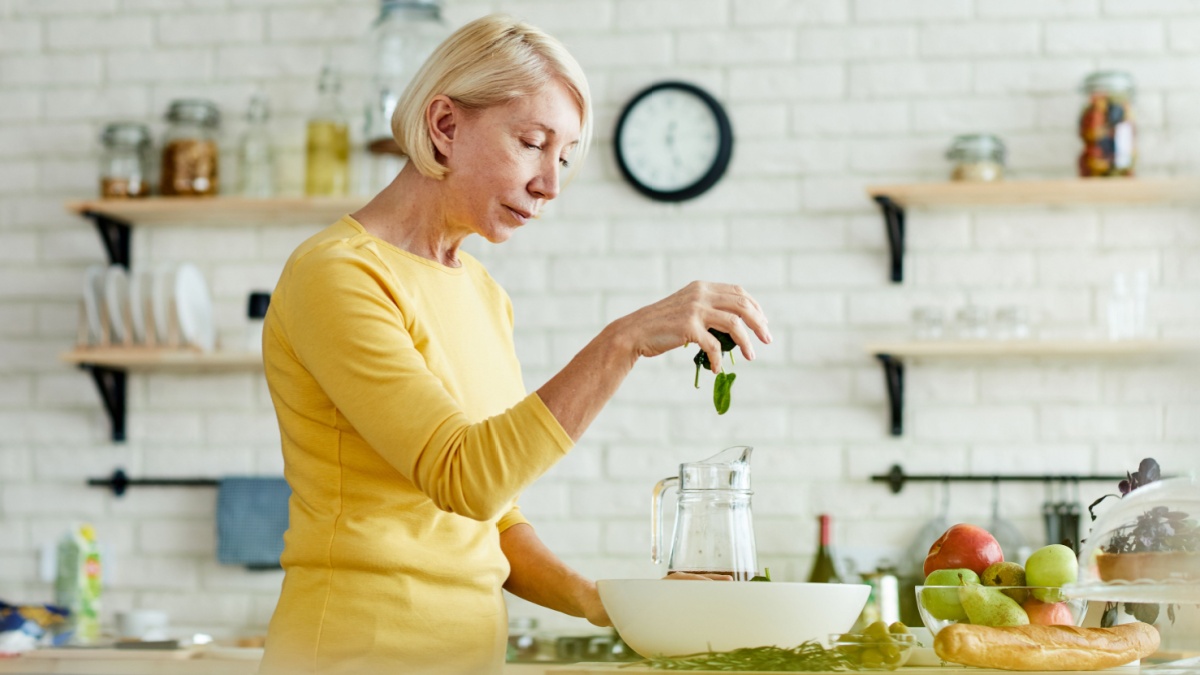 Serious attractive mature woman in yellow sweater standing at counter and adding spinach into bowl while making healthy salad in domestic kitchen.