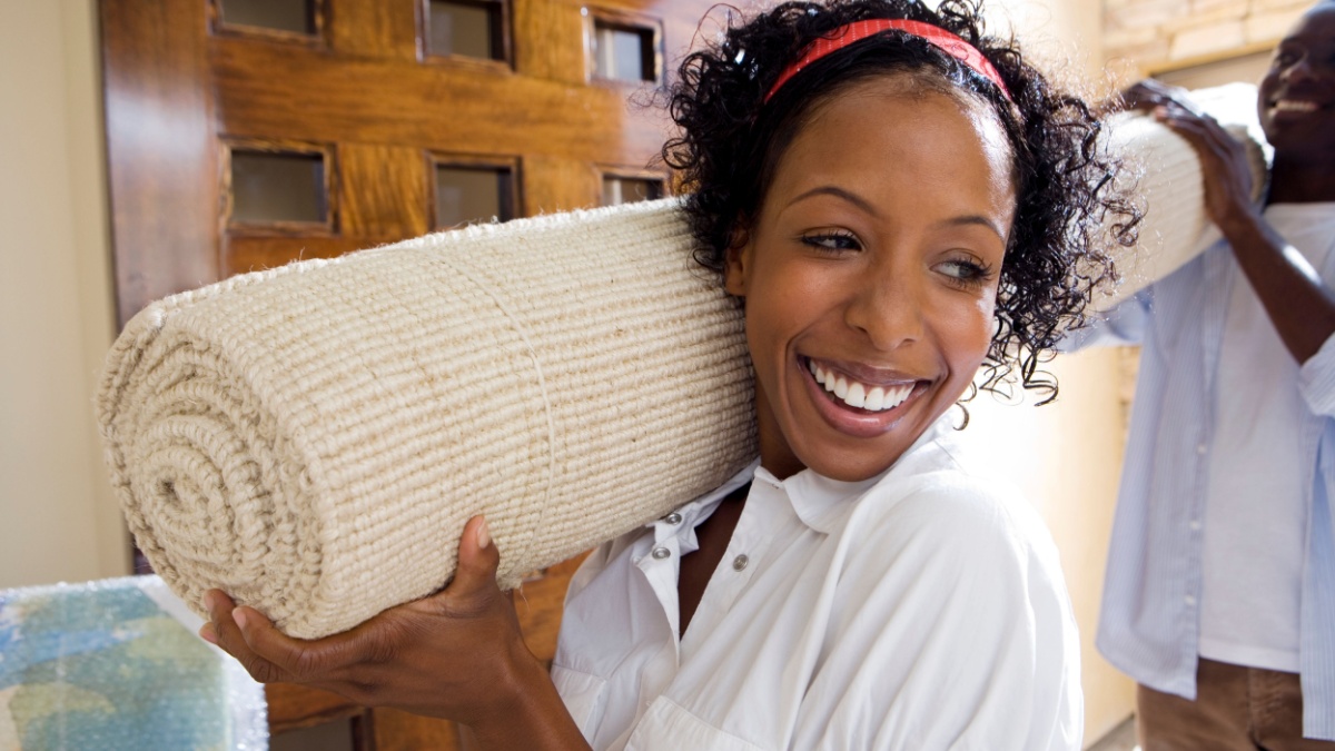 Horizontal shot of a joyous couple with the woman in front carrying a rolled up carpet on shoulder during house moving.