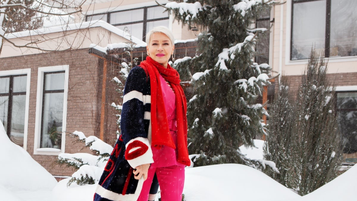 Fashionable mature woman near winter villa house, wealthy, stylish and happy aged female walking at snow near home. Short white hair Model wearing colorful trend clothes.