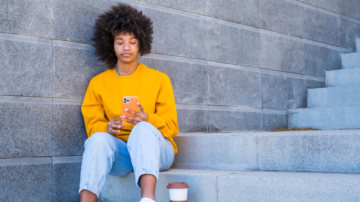 Beautiful african american woman in yellow t-shirt and curly hair using mobile phone outdoors on urban city staircase.