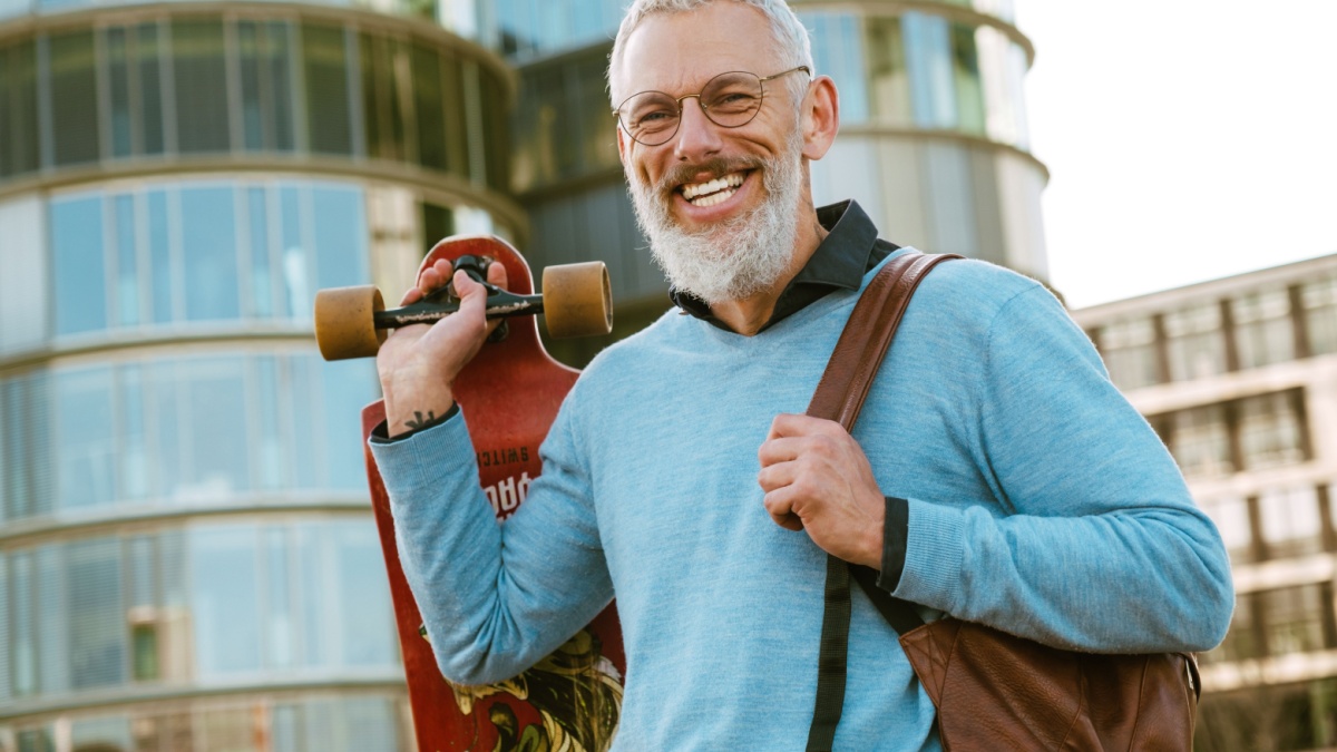 Mature grey man in eyeglasses laughing while walking on city street outdoors.