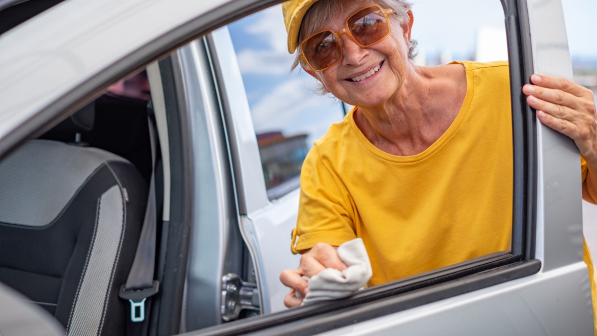 Smiling Caucasian senior woman in yellow t-shirt and hat cleans the car in a self-service car wash station taking care of the details looking at camera.