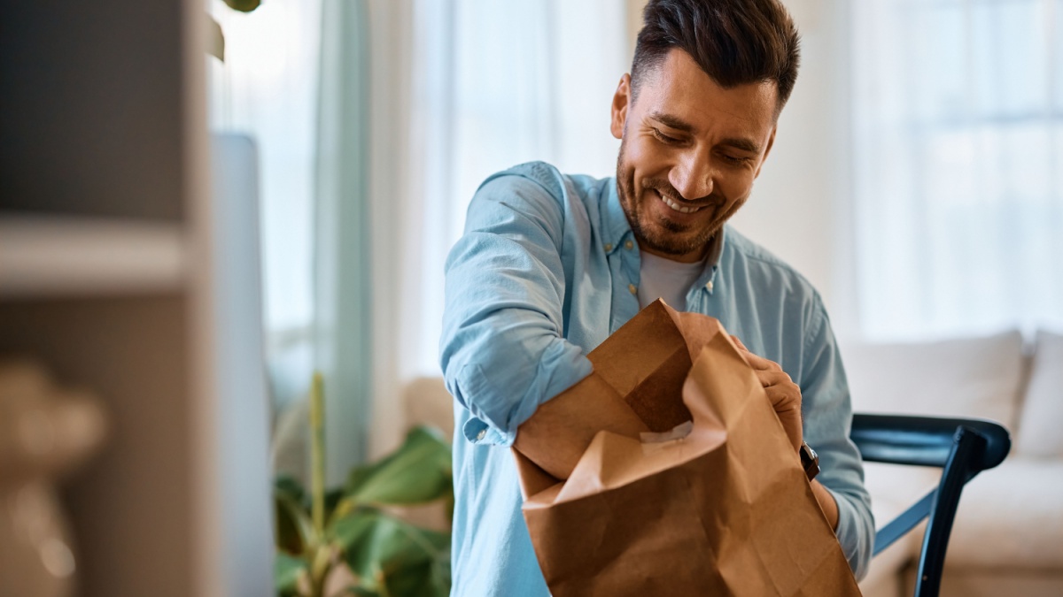 Happy man opening food delivery paper bag while working at home.