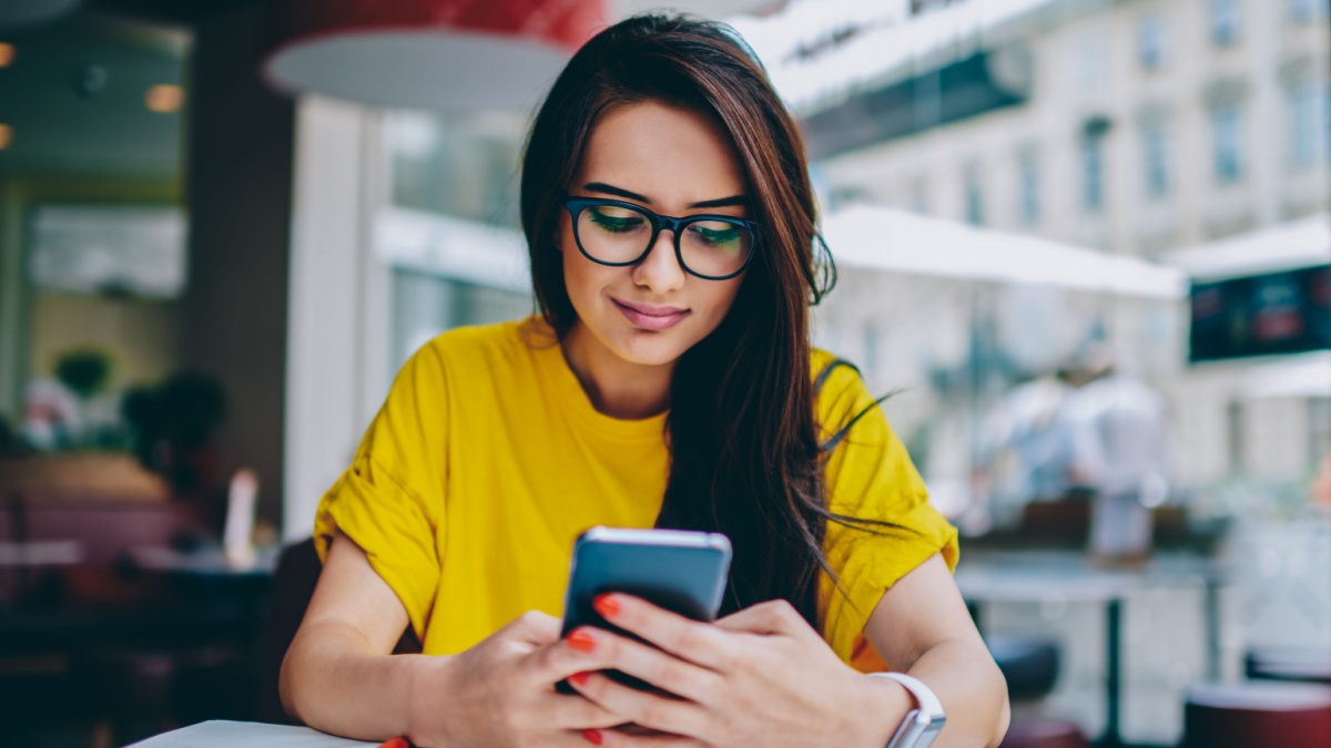 Young woman blogger in spectacles publishing new post on own website and installing new application for editing photos in social networks on digital smartphone device connected to free 4G internet.