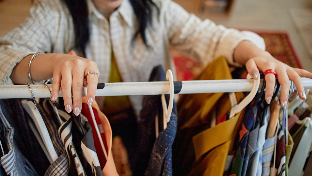 Young adult Asian woman browsing second hand clothing rack, hands holding hangers while selecting garments, partial face visible, shopping in thrift store environment.
