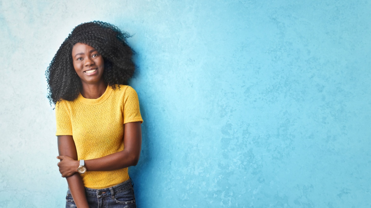 Smiling pretty Afro woman wearing yellow t-shirt standing in front of a blue wall.