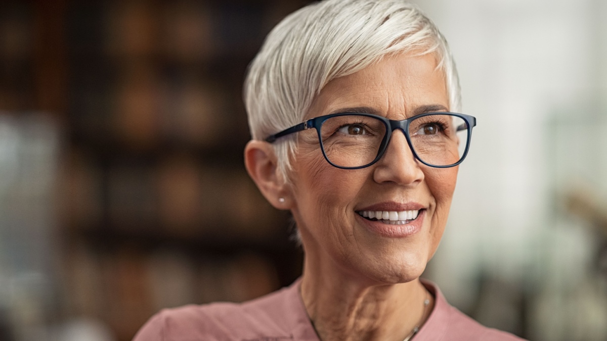 Portrait of smiling mature woman with spectacles in library looking away. Senior librarian standing in reading hall and thinking.