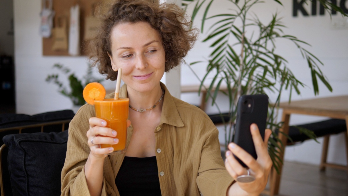 Beautiful young female blogger takes selfie pictures posing with her healthy organic cold pressed carrot juice.