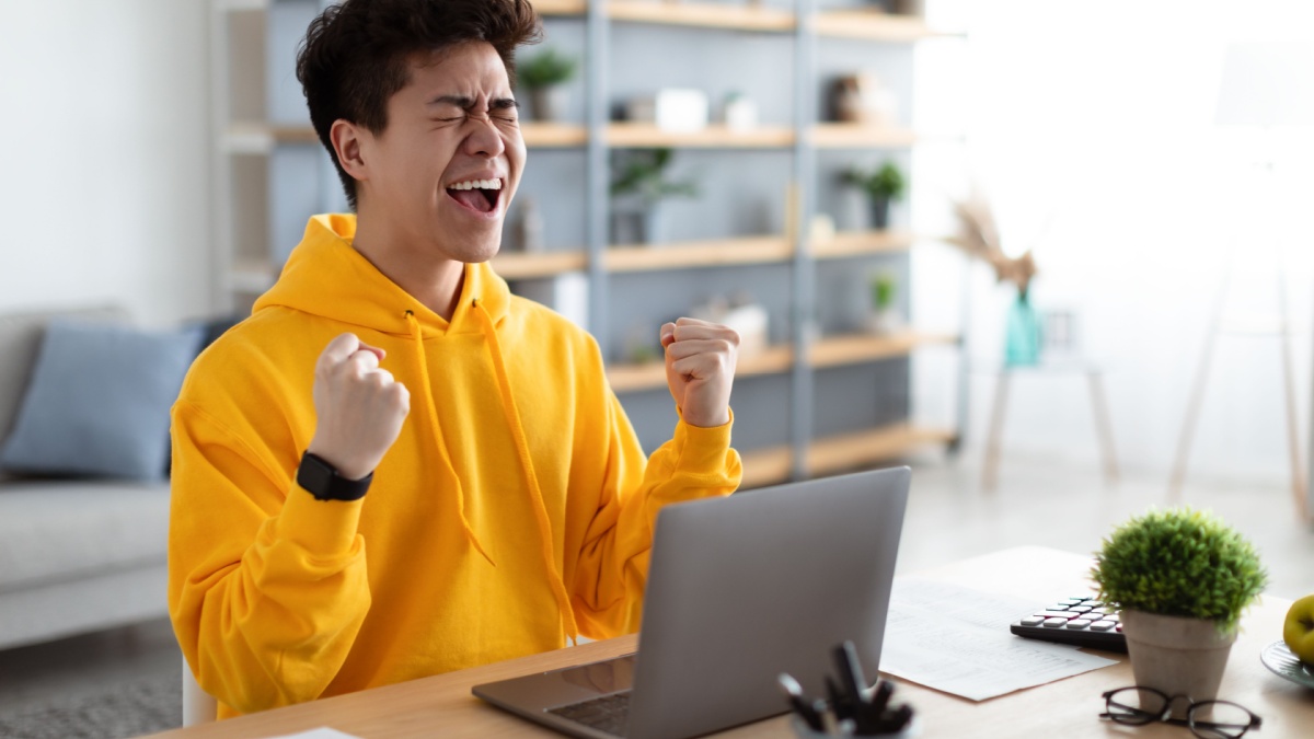 Excited asian man emotionally celebrating success using laptop in home office, sitting at desk raising hands with joy, shaking fists, received good news.