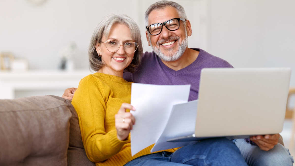 Happy senior couple holding financial documents and reading good news in letter, smiling mature family paying bills online on laptop.