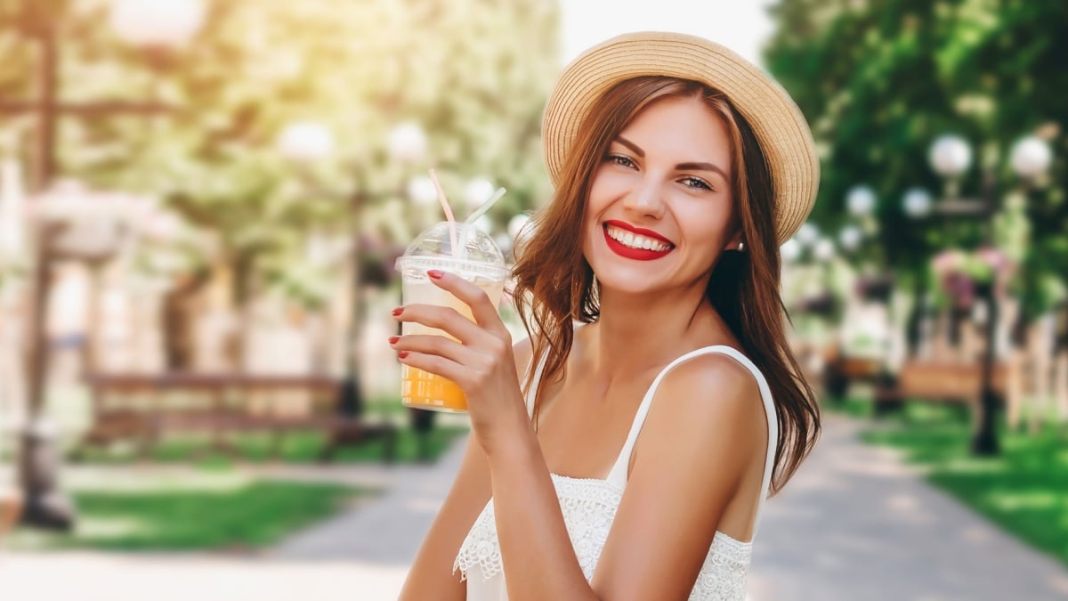 A young Ukrainian woman holds a summer refreshing cocktail and smiles against the backdrop of a public park.