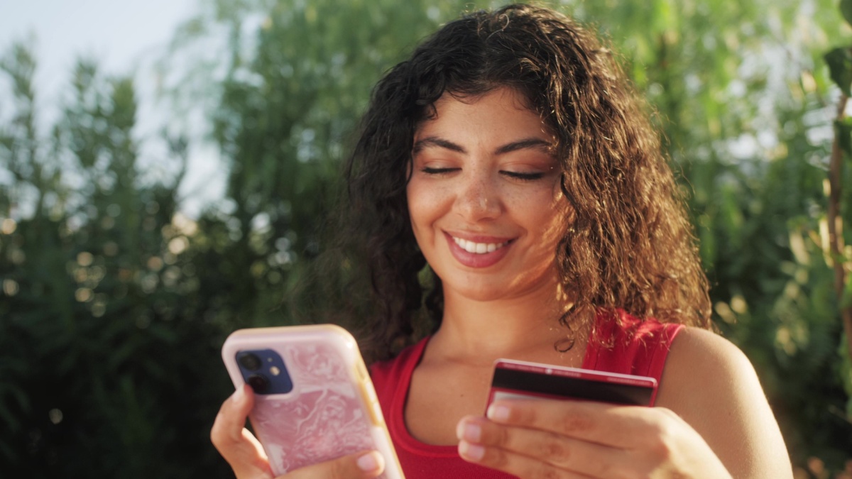 Young African American woman using cell phone holding credit card online shopping. Hispanic female shopper using instant easy mobile payments making purchase in online store. E-banking app service.