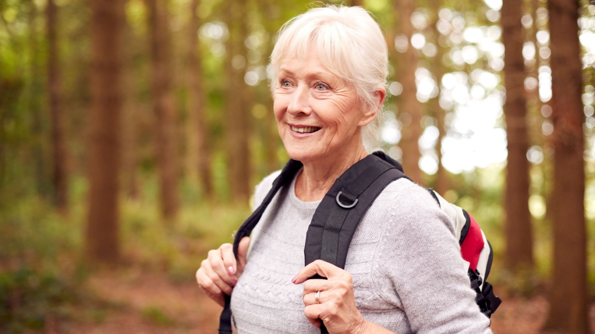 Active Retired Senior Woman Walking In Woodland Countryside Wearing Backpack.