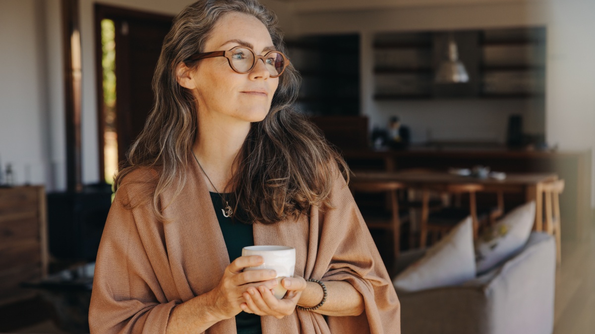 Senior woman looking away thoughtfully while standing with a cup of tea in her hands.