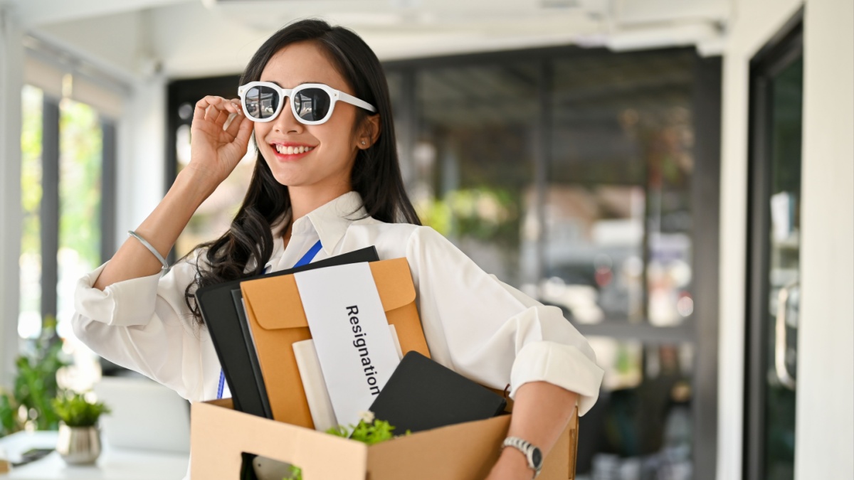 Confident and gorgeous millennial Asian female office worker, wearing sunglasses and holding a cardboard box with her belongings, feels happy to quit her job.