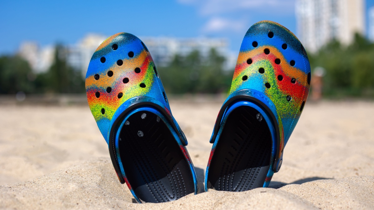 Ukraine, Kyiv - July 04. 2022: Multi-colored Crocs stick out in the sand. The beach and houses are blurred in the background. Сolorful trendy croc beach.
