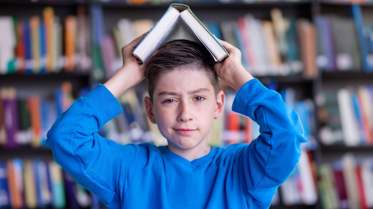 Invest in knowlenge Concept, schoolboy boy looks at the camera with a book over his head against the backdrop of the library hall.