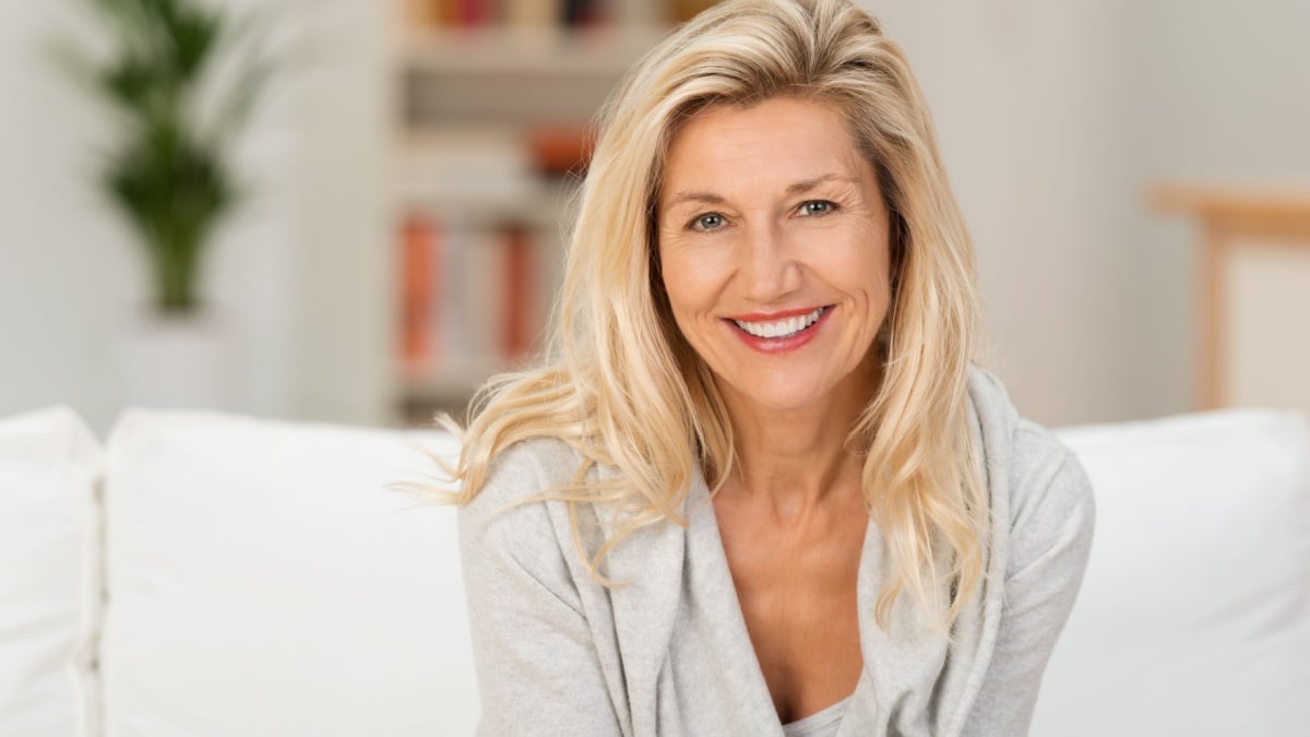 Lovely middle-aged blond woman with a beaming smile sitting on a sofa at home looking at the camera.