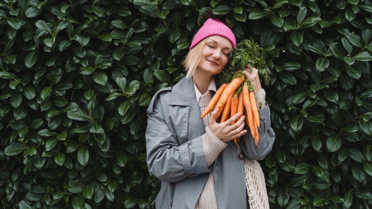 Smiling woman with fresh organic carrots and mesh bag against green leaf wall. Concept of people doing sustainable shopping of eco local products without plastic packaging. Minimalist lifestyle.
