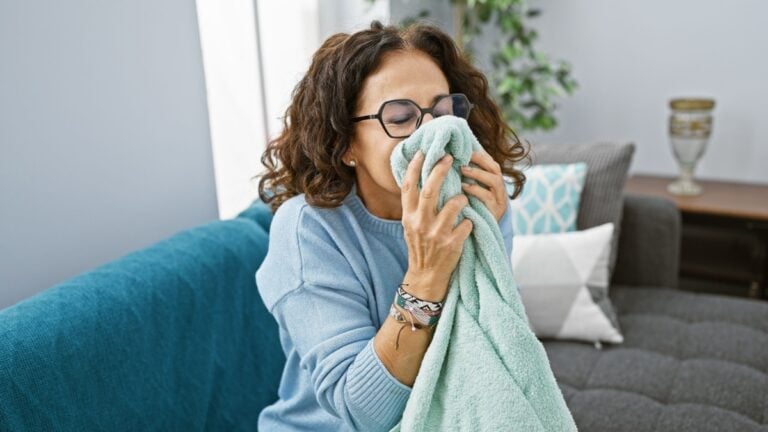 A woman smiles while holding a towel to her face in a cozy living room, illustrating relaxation and domestic life.