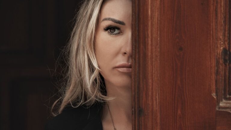 Beautiful girl peeks through the door, peeking Out of Doorway. GIrl with low lighting, a massive door covers half face closeup. Suspicious and attentive look.