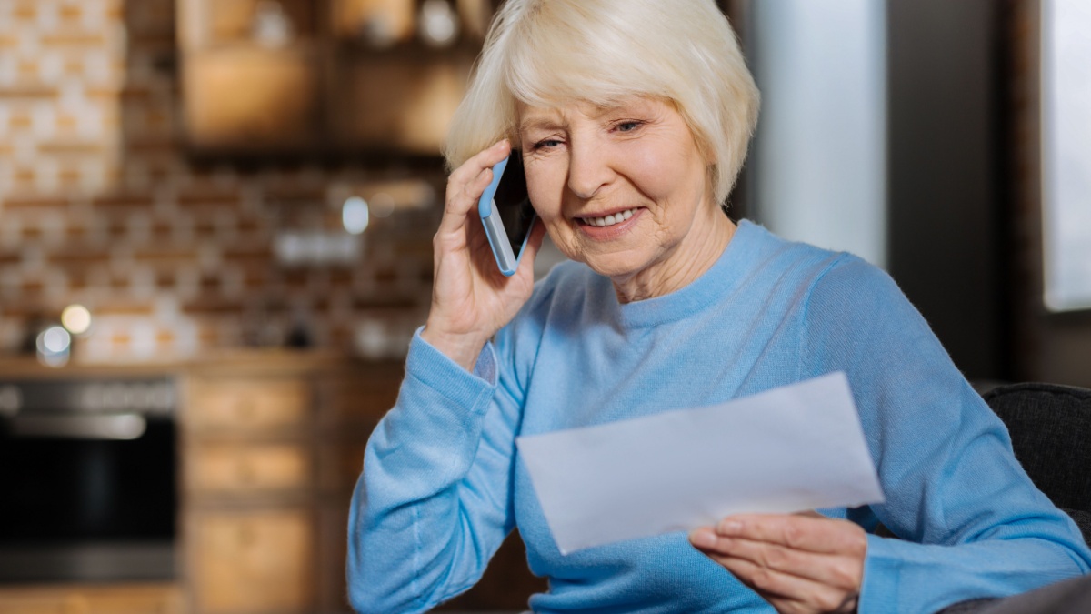 Distance communication. Delighted positive elderly woman holding a bill and smiling while making a phone call.