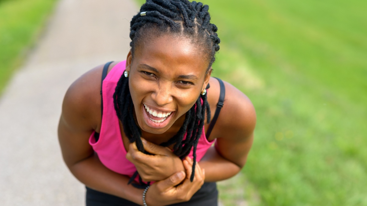 Sporty young African woman bending forwards laughing at the camera as she takes a break from jogging on a rural footpath.
