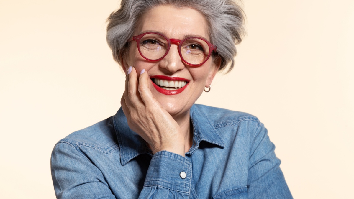 Close up portrait of attractive senior woman in modern red eyeglasses. Happy older female teacher with trendy haircut and toothy smile on beige background. Eyesight problem and stylish eye.