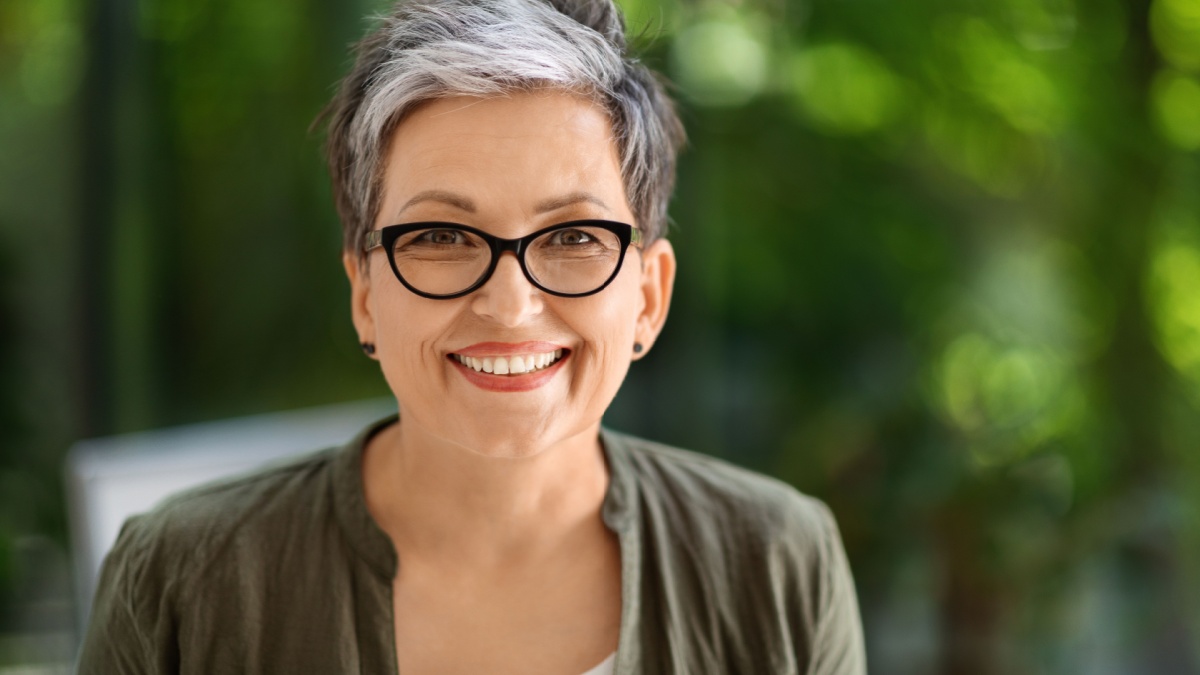 Portrait of happy attractive short-haired stylish middle aged 50s businesswoman wearing eyeglasses smiling at camera, posing at green eco-friendly office.