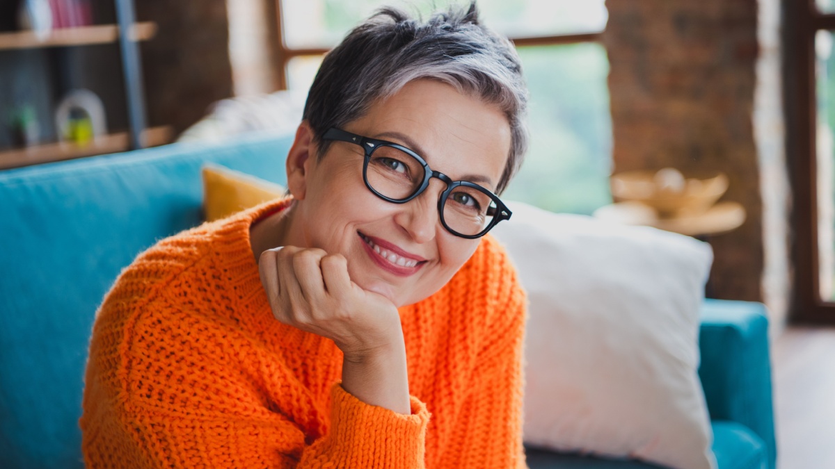 Photo of positive dreamy elderly lady wear orange pullover spectacles arm hand chin sitting sofa indoors apartment room.