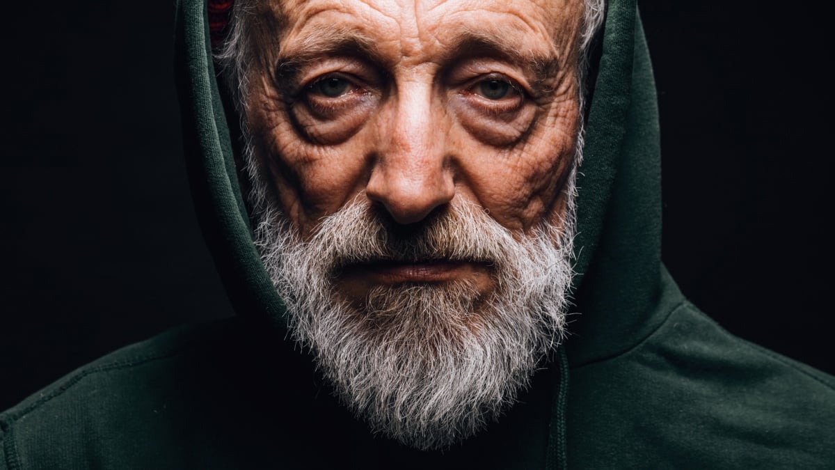 Close-up of a mature old-aged man in green hoodie with wrinkled weathered face, puffy sad eyes looking at camera with mournful expression.