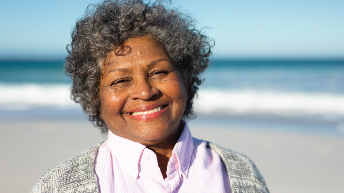 Portrait close up of a senior African American woman standing on the beach with blue sky and sea in the background, smiling to camera.