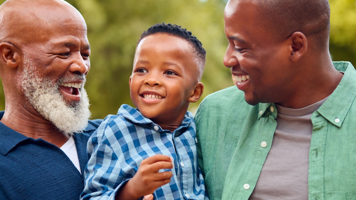 Three Generation Male Family Laughing Hugging Grandson Standing Outdoors In Countryside.