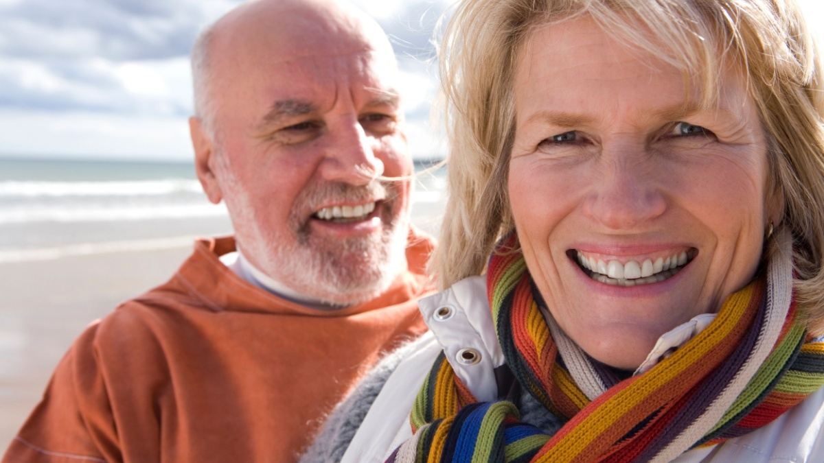 Active senior couple walking on beach by sea in autumn at camera.