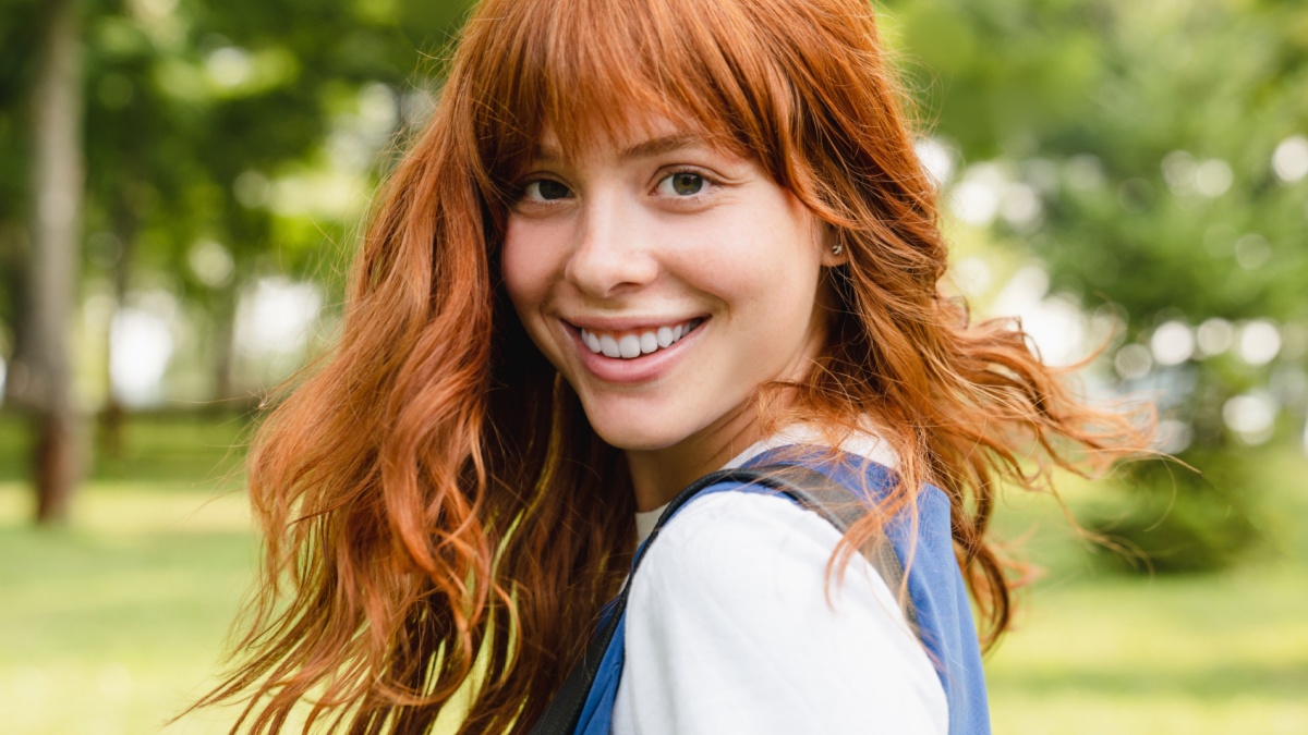 Close up portrait of a young caucasian woman girl teenager student schoolgirl with ginger red hair and toothy smile walking in park forest outdoors looking at camera.