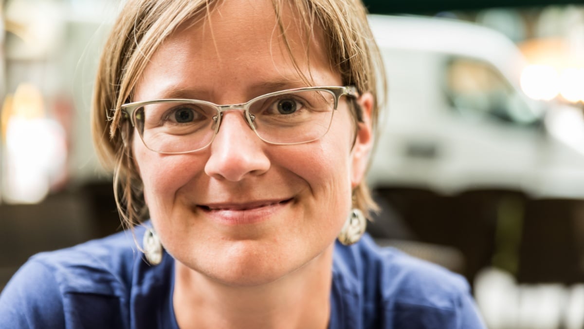Close up portrait of a smiling young white woman with glasses wearing a blue and sweater with a bokeh city background.