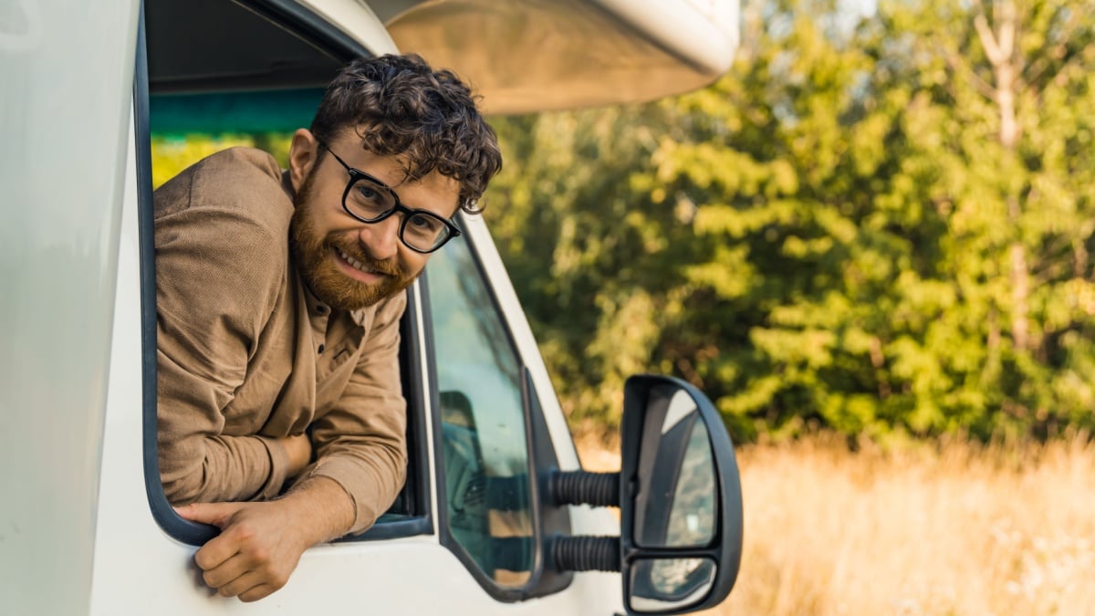 Cheerful young bearded man leaning out of the window of a camper van and smiling at the camera.