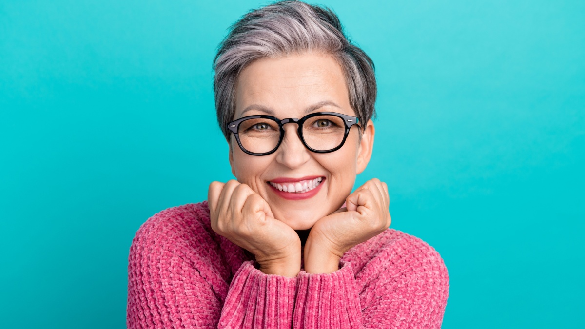 Photo of good mood woman with short hair dressed pink sweater in glasses holding finsts under chin isolated on teal color background.