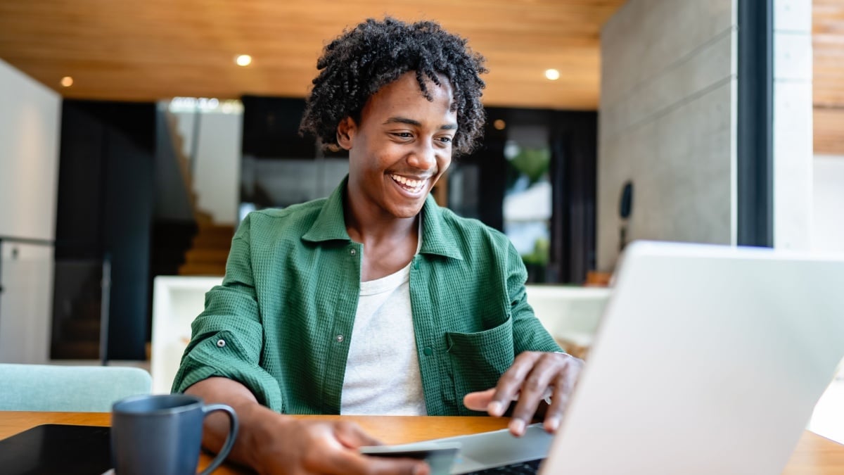 A young man of African descent sits by a wooden table in a modern, sunlit living room, smiling as he uses a laptop to make an online purchase and pays by credit card.