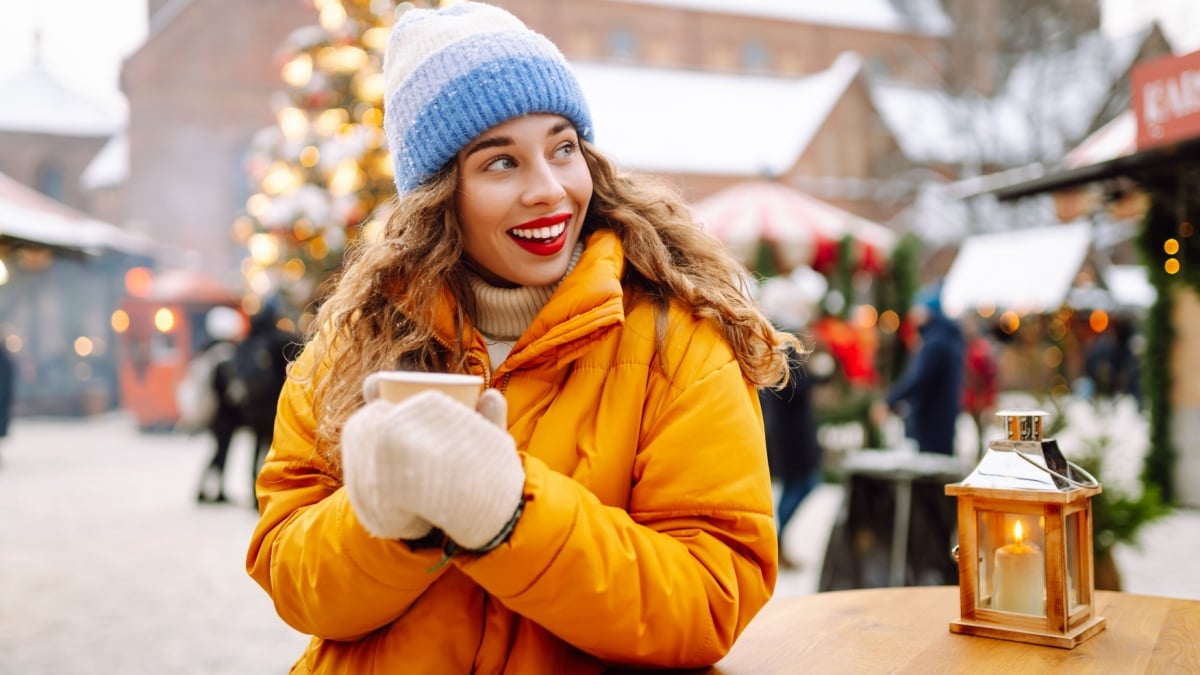 Happy woman drinks a hot drink at a Christmas market decorated with festive lights.
