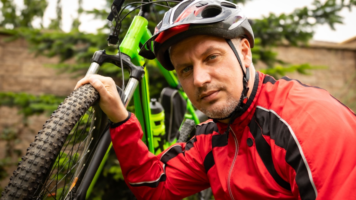 A man in a red jacket and helmet holding a green bicycle with a serious expression, outdoors on a lawn with a blurred background.